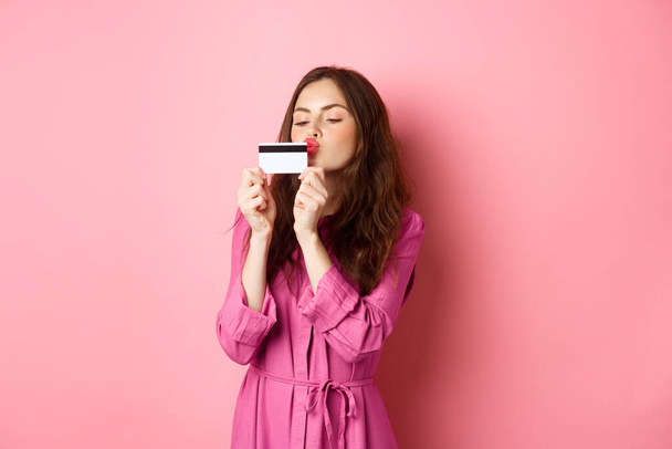 Изображение молодой красивой женщины, целующей свою кредитку, идущей по магазинам, тратящей деньги в магазинах, стоящей на розовом фоне - Фото, изображение