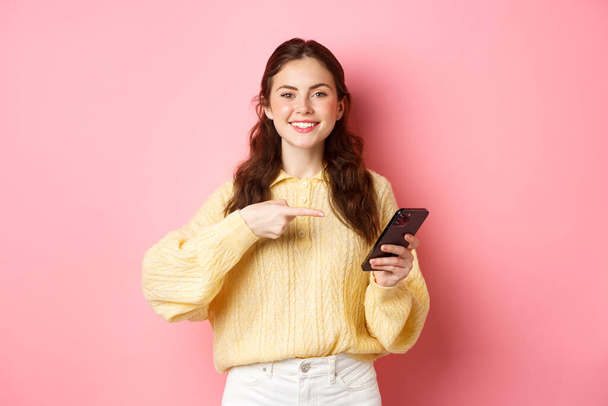 Image of happy, smiling young woman, holding mobile phone and pointing at mobile screen with pleased and confident face, standing over pink background - Photo, Image