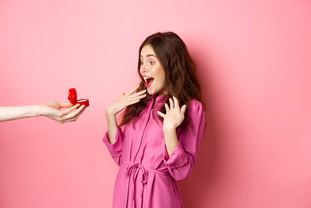 Porträt der überraschten Freundin erhalten Heiratsantrag, Blick auf die Hand mit Verlobungsring und Schrei der Überraschung und Aufregung, steht über rosa Hintergrund - Foto, Bild