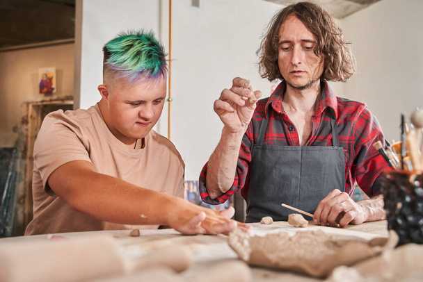 Man with special needs working with clay in ceramic studio under supervision of craftsman - Zdjęcie, obraz