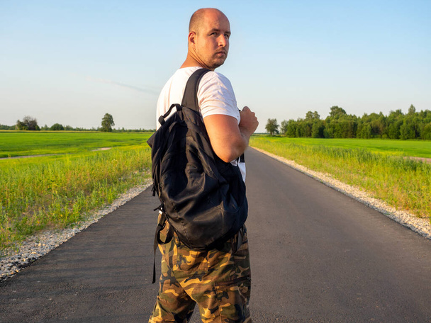 Ένας άντρας στέκεται σε έναν ασφαλτοστρωμένο δρόμο με ένα μαύρο σακίδιο και κοιτάζει πίσω του. Η έννοια του ταξιδιού και της περιπέτειας - Φωτογραφία, εικόνα