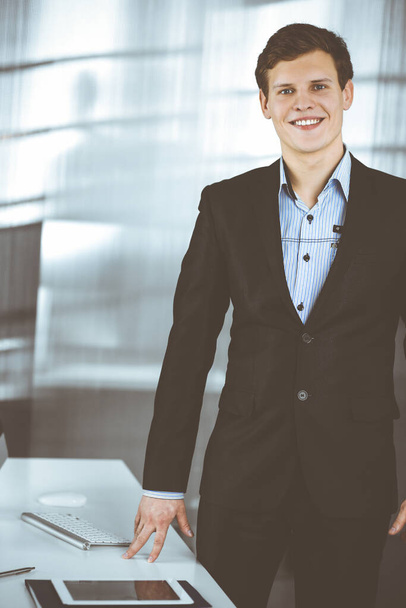 Friendly successful businessman in a black suit is standing at his workplace. Portrait of a young specialist in a modern office. Business concept - Фото, изображение