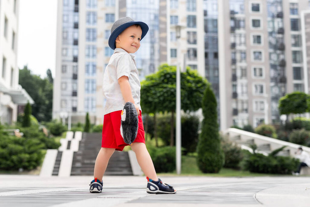 Cute adorable caucasian 3-4 years blond little kid boy in stylish shorts shirt and hat hold soft teddy toy enjoy playing outdoors at urban city center downtown area. Funny child portrait - Photo, Image