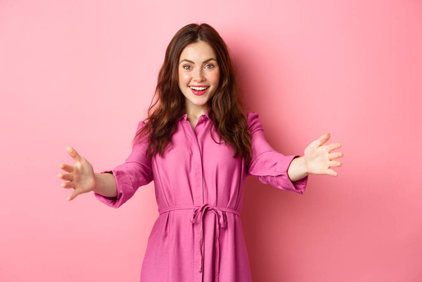 Стильная женщина, протягивающая руки, чтобы поприветствовать друга, хочет обнять или пригласить кого-то, стоя на розовом фоне - Фото, изображение