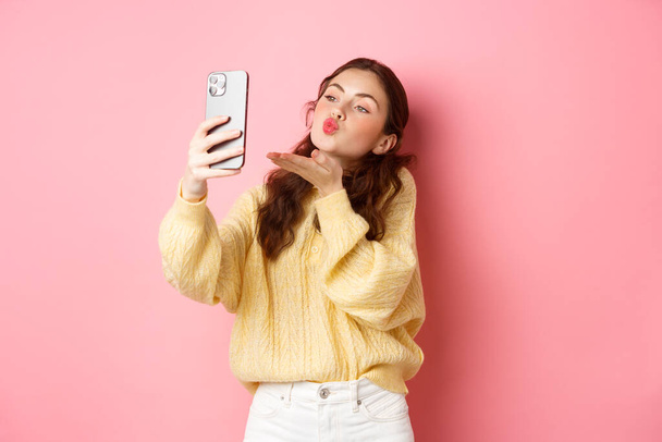 Beautiful flirty girl taking selfie on smartphone, sending air kiss during video chat, standing against pink background - Foto, Imagem
