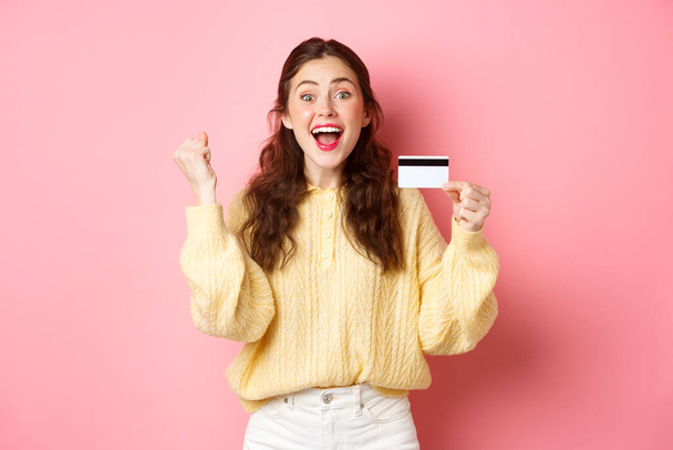 Ενθουσιασμένη νεαρή γυναίκα κερδίζει βραβείο, δείτε φοβερό πώληση promo, δείχνοντας πλαστική πιστωτική κάρτα της και κραυγή της χαράς, λέγοντας ναι, στέκεται σε ροζ φόντο - Φωτογραφία, εικόνα