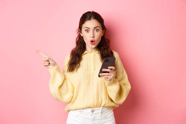 Έκπληκτη νεαρή όμορφη κοπέλα λαχανιάζει και λέει wow, δείχνοντας δάχτυλο στην άκρη αριστερά copyspace, κρατώντας το κινητό τηλέφωνο, στέκεται πάνω σε ροζ φόντο - Φωτογραφία, εικόνα