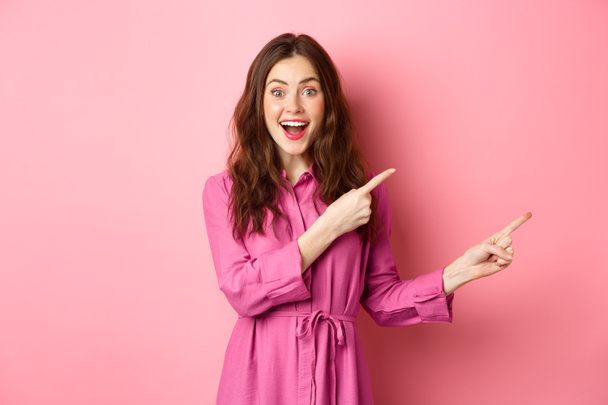 Belleza y moda. Sorprendida mujer moderna elegante señalando los dedos a la derecha, mostrando promo en copyspace a un lado, de pie sobre fondo rosa - Foto, imagen