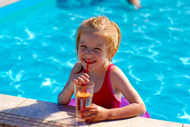 Happy little blonde girl with a pink swimming circle drinks lemonade through a straw in the pool. - Foto, imagen