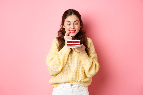 Chica linda tentada mira sabrosa torta, quiere tener un bocado de postre, sosteniendo el dedo en los labios y mirando fijamente al plato, de pie sobre el fondo rosa - Foto, imagen