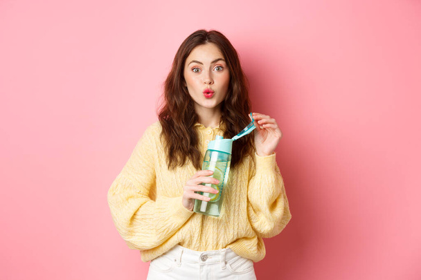 Portret van een opgewonden brunette die water drinkt met citroen uit een sportfles, kijk geïntrigeerd en geïnteresseerd op de camera, staande tegen een roze achtergrond - Foto, afbeelding