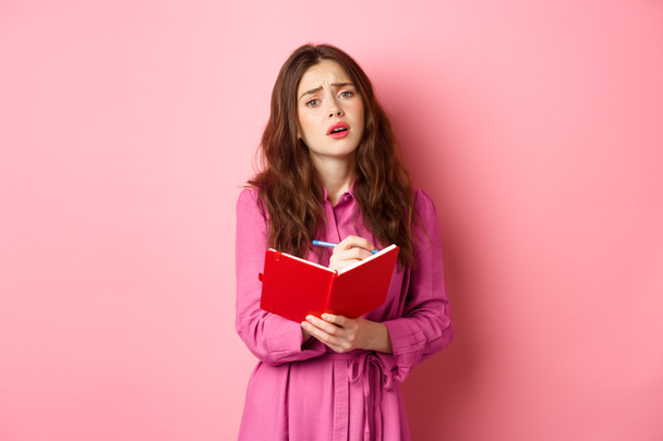 Скромная романтичная девушка вздыхает, пишет в своем дневнике о печальном моменте, делает заметки в планировщике, стоя на розовом фоне - Фото, изображение