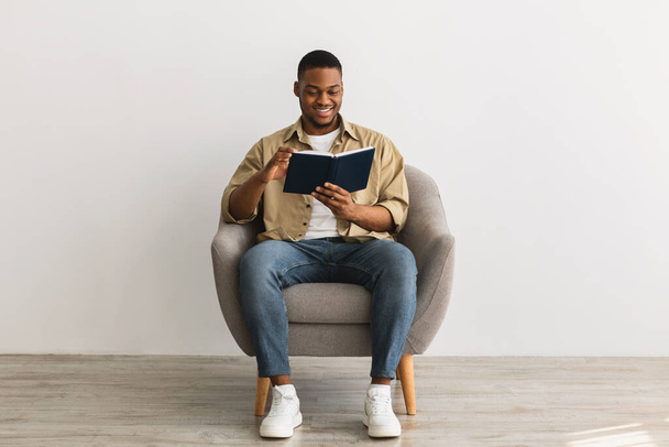 zwart man lezen boek zitten in fauteuil op grijze achtergrond - Foto, afbeelding