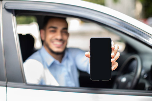 Wesoły arabski mężczyzna siedzący w samochodzie, pokazujący komórkę, makietę - Zdjęcie, obraz