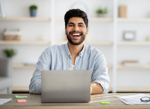 Retrato del exitoso freelancer árabe sentado en el lugar de trabajo con computadora portátil y sonriendo a la cámara, espacio para copiar - Foto, Imagen