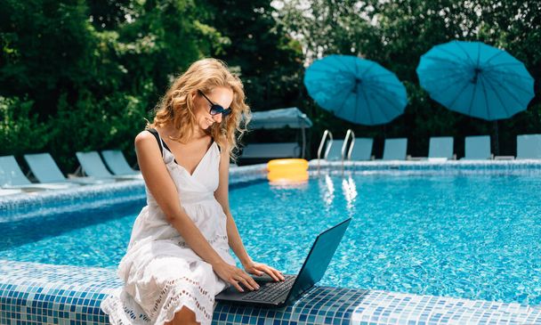 Focused female freelancer with laptop remote worker sitting near pool at hotel. Recreational work and health. Summer or winter tourism concept. Blue clear water in the pool. High quality photo - Photo, Image