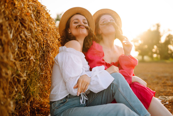 Two young woman in fashionable and stylish clothes posing near hay bales in the countryside. Fashion concept. Nature, vacation, relax and lifestyle. - Photo, image