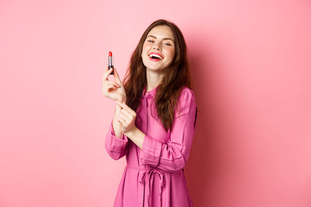 Beautiful woman smiling, applying lipstick and make up for party, standing against pink background, getting ready for going out - Photo, Image