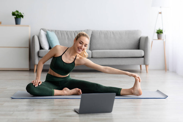 Νεαρή ξανθιά αθλητική γυναίκα στα αθλητικά κάνει προπόνηση στο πάτωμα στο σπίτι και βλέποντας βίντεο γυμναστικής στο laptop - Φωτογραφία, εικόνα