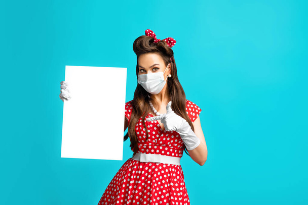 Joven pinup mujer en la máscara anti-covid apuntando a cartel de papel en blanco con maqueta para su diseño sobre fondo azul - Foto, imagen