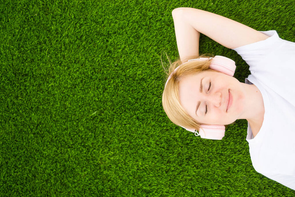 Une jeune fille caucasienne en t-shirt blanc et écouteurs se trouve sur une pelouse artificielle avec les yeux fermés. Le concept de l'écoute de la musique, podcasts. Minimalisme, espace de copie, vue de dessus. - Photo, image