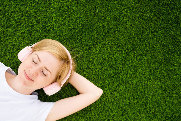 Młoda biała dziewczyna w białej koszulce i słuchawkach leży na sztucznym trawniku z zamkniętymi oczami. Koncepcja słuchania muzyki, podcastów. Minimalizm, przestrzeń do kopiowania, widok z góry. - Zdjęcie, obraz