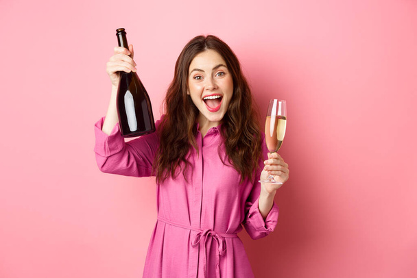 Feest- en feestdagen concept. Gelukkige vrouw dansen en plezier hebben, champagne drinken, fles en glas vasthouden, toast maken, over roze achtergrond staan - Foto, afbeelding