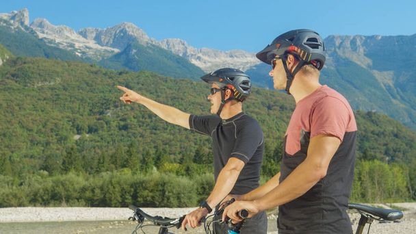 CLOSE UP: Zwei Freunde auf Mountainbikes schauen sich um und zeigen auf die Landschaft. - Foto, Bild