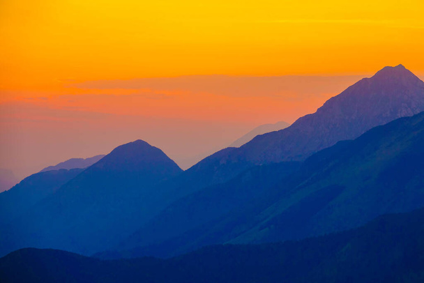AERIAL: Burnt orange evening sky spans above the blue hued mountain range. - Photo, Image