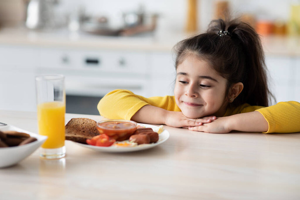 Hungriges kleines Mädchen lehnt am Tisch und blickt auf Teller mit leckerem Essen - Foto, Bild