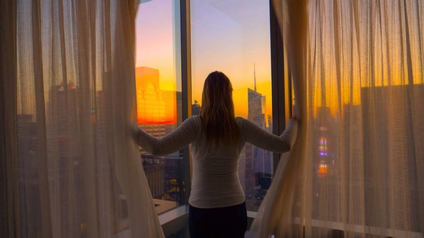 閉じる:認識できない女性がカーテンを開き、日没時に街を見る. - 写真・画像