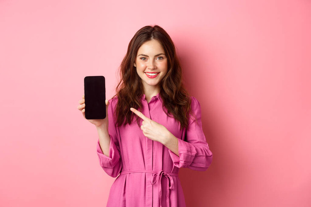 Nuori valkoihoinen nainen tyylikäs mekko, sormella älypuhelimen näytön ja hymyillen, osoittaa promo paljon verkossa, vaaleanpunainen tausta - Valokuva, kuva
