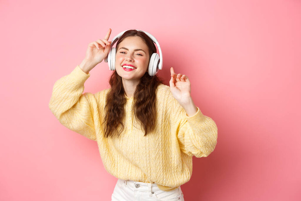 Mooi stijlvol vrouwelijk model zingen favoriete lied, luisteren naar muziek in draadloze hoofdtelefoon, glimlachen en dansen, staande over roze achtergrond - Foto, afbeelding