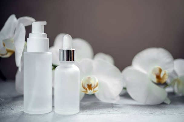 White glass serum bottle, lotion pump bottle and cream jar mock-ups in the bathroom with orchid flowers in the background, unbranded cosmetic products, spa cosmetic product branding mockup - Photo, Image