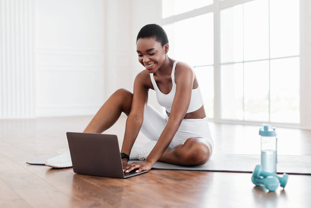Souriant jeune femme noire assise sur un tapis de yoga à l'aide d'un ordinateur portable - Photo, image