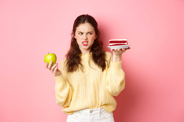 Gesundes Mädchen auf Diät schaut angewidert auf ein Stück Kuchen, hält grüne Applikation in der Hand, grimmig unzufrieden, steht vor rosa Hintergrund - Foto, Bild