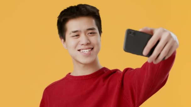 Chico asiático haciendo selfie en Smartphone posando sobre fondo amarillo - Imágenes, Vídeo
