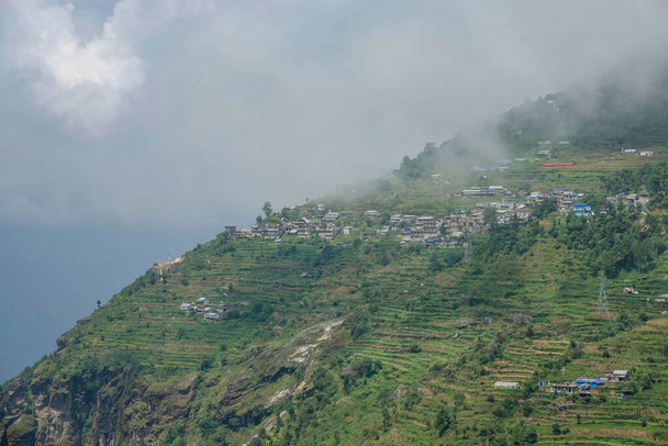 AERIAL: White clouds drifts over the rural village on the steep hill in Nepal. - 写真・画像