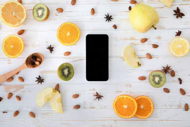 健康的な食事の概念。果物のスライス、アーモンドとスターアニス、トップビューと木製のテーブルの上に空白の黒い画面を持つスマートフォン - 写真・画像