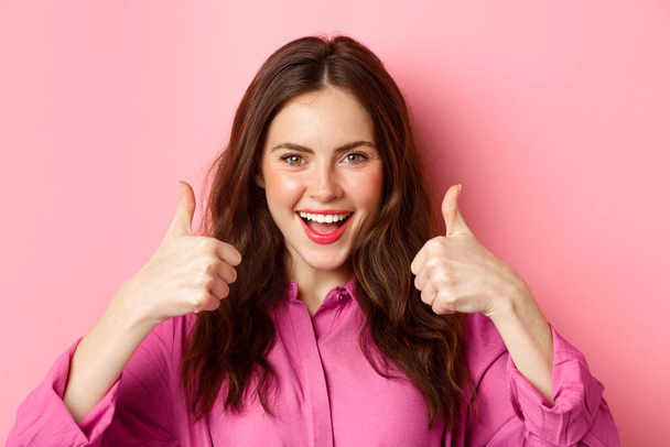 Confident attractive woman say yes, shows thumbs up with determined face, praise you, nice work gesture, standing over pink background - Foto, afbeelding