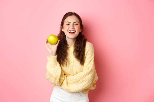 Здоровое питание, люди и образ жизни. Яблочный день отпугивает доктора, девушка держит вкусные фрукты и улыбается счастливо перед камерой, стоя на розовом фоне - Фото, изображение