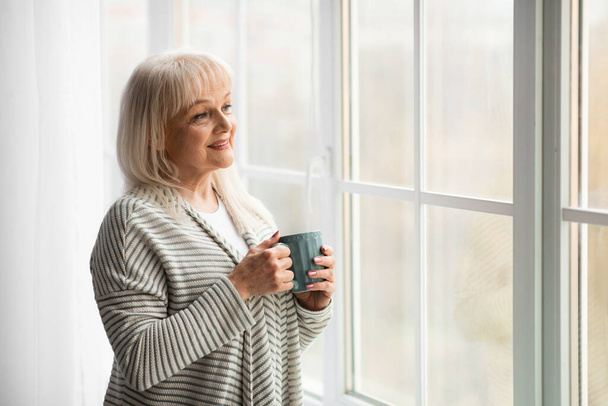 Mature woman drinking hot coffee looking out of window - Фото, изображение