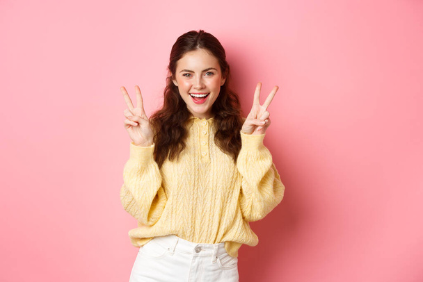 Portrait d'une jolie mannequin brune féminine, souriante et montrant des signes de paix, faisant un geste de signe V, debout positif et heureux sur fond rose - Photo, image
