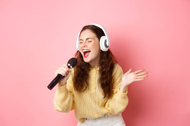 Πορτρέτο της νεαρής γυναίκας τραγουδώντας καραόκε, φορώντας ακουστικά και ερμηνεύοντας το τραγούδι, κρατώντας μικρόφωνο, στέκεται σε ροζ φόντο - Φωτογραφία, εικόνα