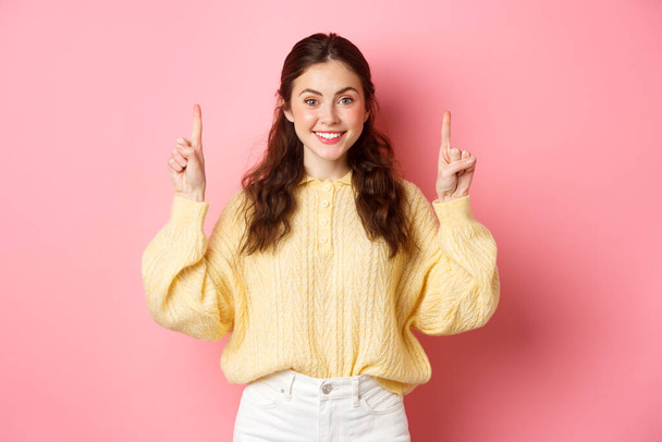 Image of attractive young caucasian woman pointing fingers up, showing advertisement, smiling at camera, standing against pink background - Foto, afbeelding