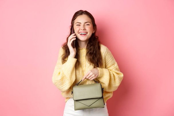 Roztomilá běloška mluví do telefonu a směje se, drží kabelku, šťastně se usmívá do kamery, stojí na růžovém pozadí - Fotografie, Obrázek
