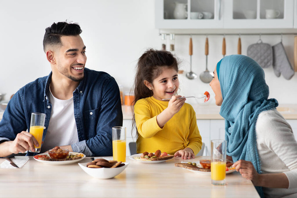 Маленькая арабская девочка кормит маму во время завтрака с родителями на кухне - Фото, изображение