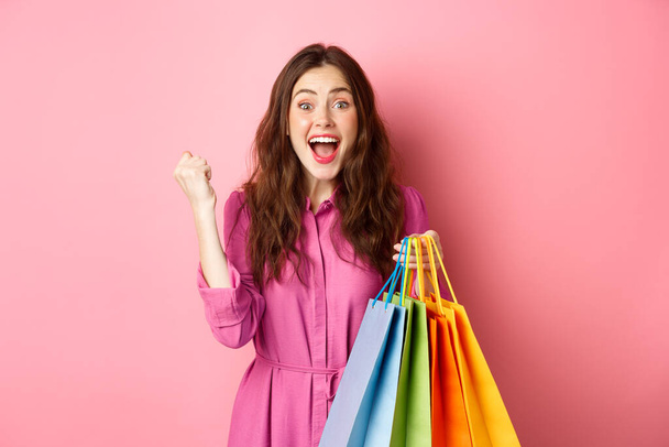Возбужденная девушка кричит от радости, делает кулачный насос, держа сумку с покупками и радуясь, стоя в платье на розовом фоне - Фото, изображение