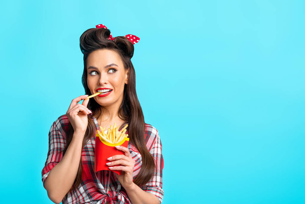 Young pinup woman in retro outfit eating french fries from package, looking aside at empty space on blue background - Photo, image