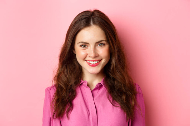 Close up retrato de mulher moderna elegante com maquiagem e penteado encaracolado, sorrindo e olhando determinado para a câmera, de pé contra o fundo rosa - Foto, Imagem